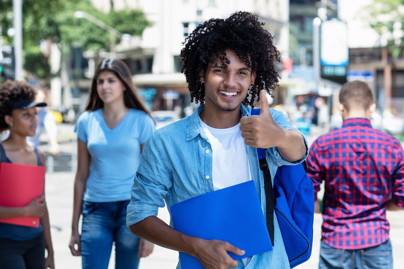 Laughing mexican male student showing thumb up with other young adults outdoor in summer in city